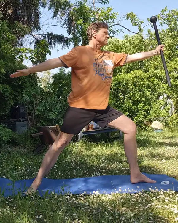 Werner Hauschild im Krieger II auf einer Yogamatte im Garten, den Steel Mace in der ausgestreckten Hand