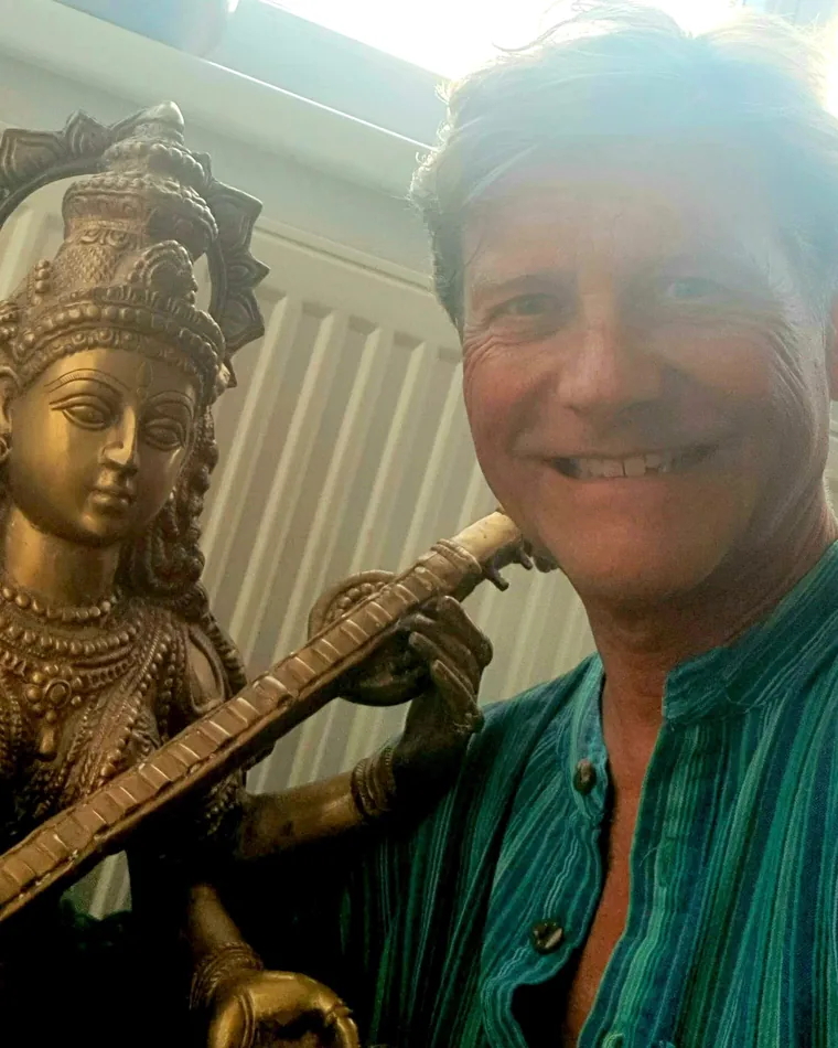 Werner Hauschild lacht in die Kamera, neben ihm eine Messingfigur der Göttin Saraswati mit Vina